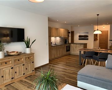 Ein modernes Wohnzimmer mit hellem Holz-Mobiliar und einer offenen Küche. Die Einrichtung ist gemütlich und einladend, mit Pflanzen und warmem Licht.