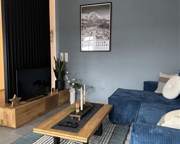 Ein modernes Wohnzimmer mit einem blauen Sofa und einem Holztisch. An der Wand hängt ein Bild und eine schwarze Struktur hebt den Raum hervor.