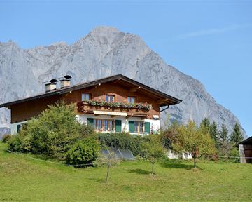 Ein typisches Alpenhaus mit Balkon und Blumen. Im Hintergrund erheben sich majestätische Berge unter einem klaren Himmel.