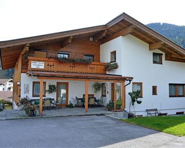 Ein schönes Holzhaus mit einem Balkon und einem begrünten Bereich. Im Hintergrund sind sanfte Hügel zu sehen.